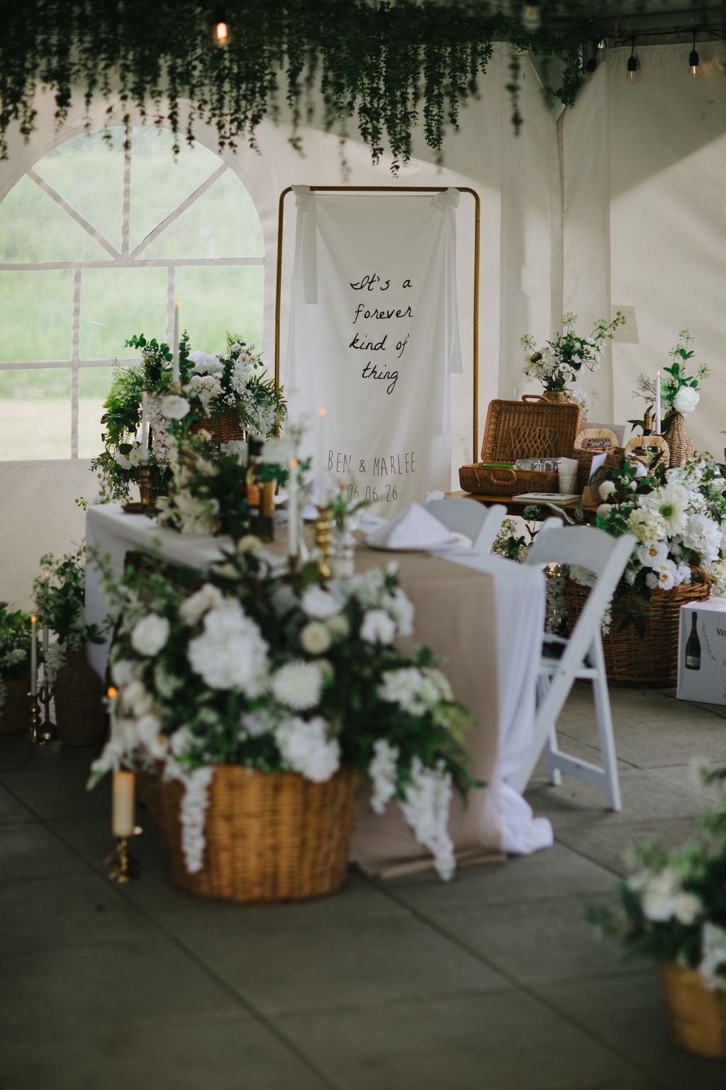 Elegant wedding ceremony backdrop with handwritten sign reading "it''s a forever kind of thing" surrounded by white flowers in wicker baskets and cascading gr…