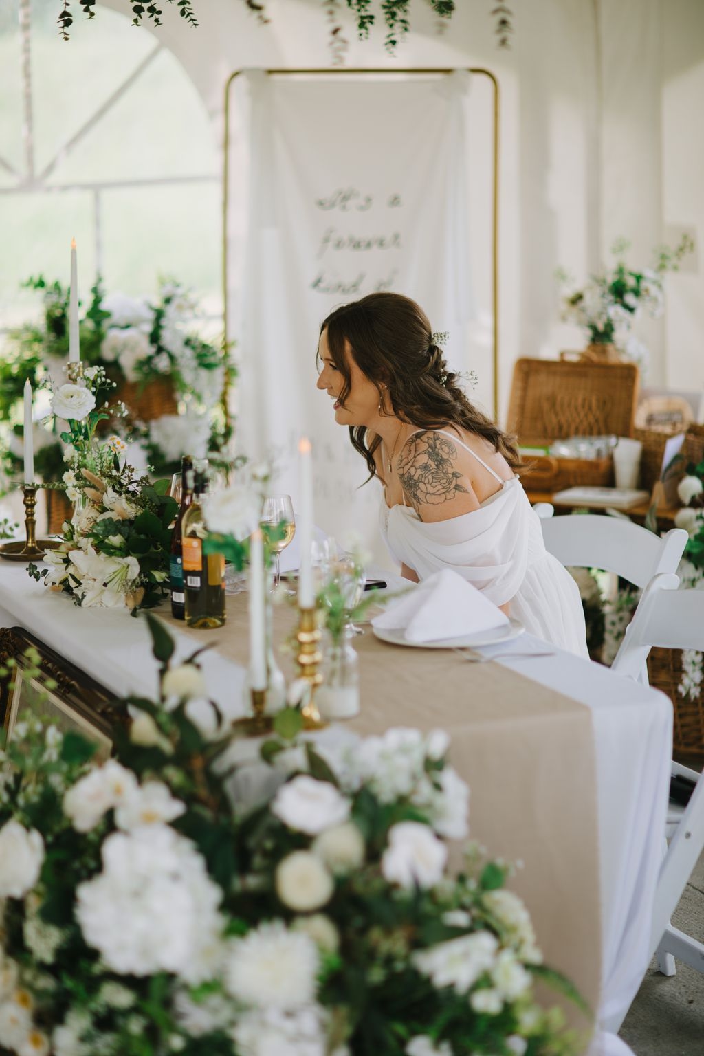Bride in white off-shoulder dress seated at elegant table with white flowers, greenery, and candlelight, gazing thoughtfully to the side