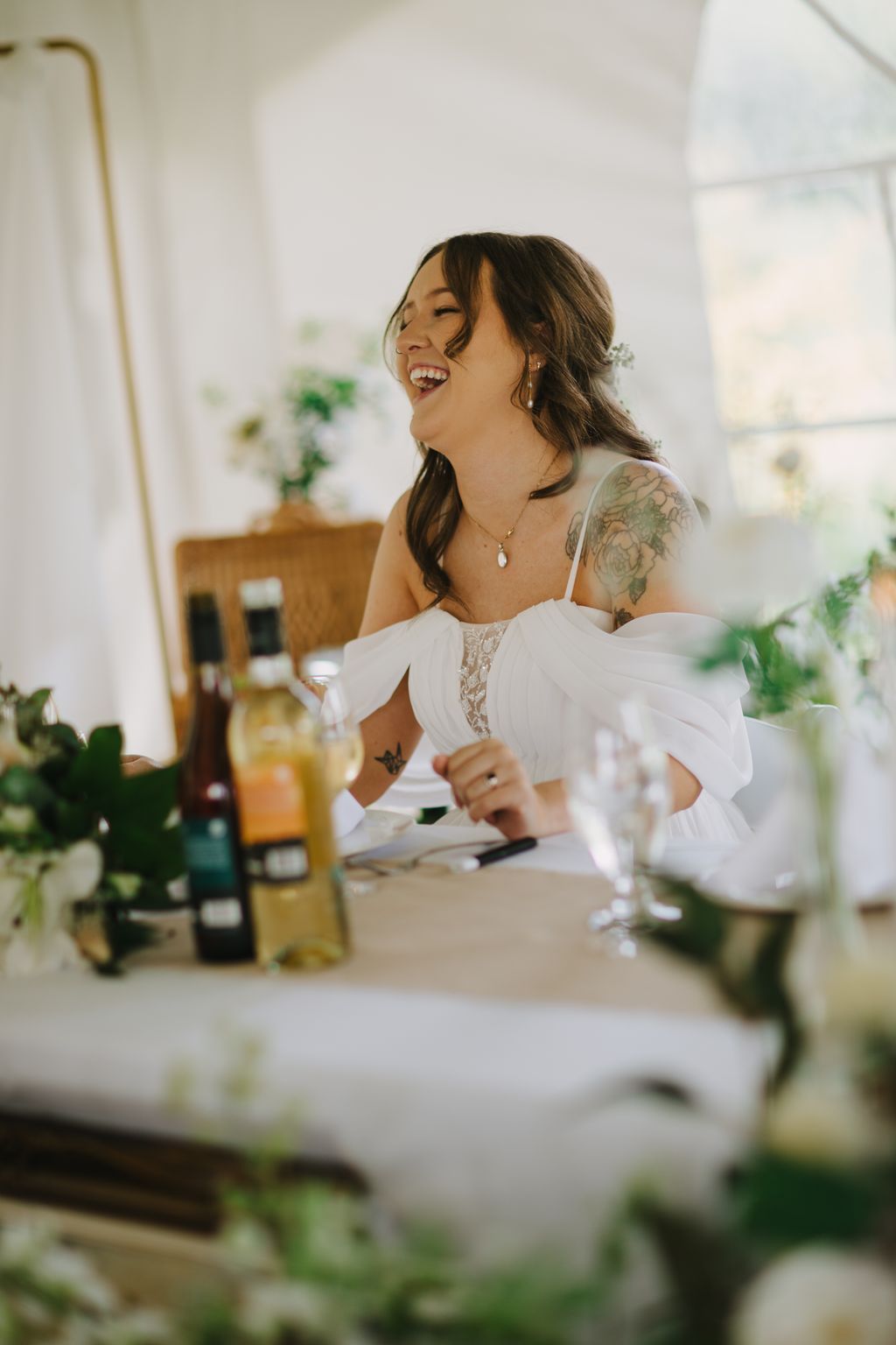 Bride in white off-shoulder lace dress laughing joyfully at vanity table with soft natural light and blurred green foliage