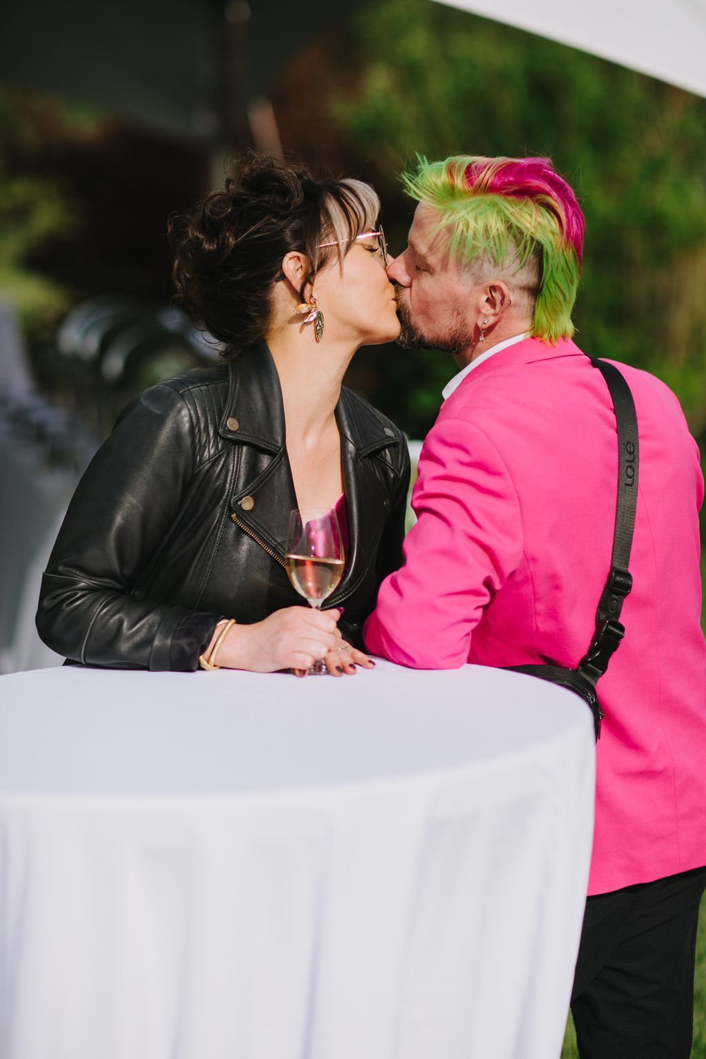 Couple kissing outdoors in bright daylight, bride in black leather jacket holding champagne flute, groom in vibrant pink jacket with neon green and pink hair