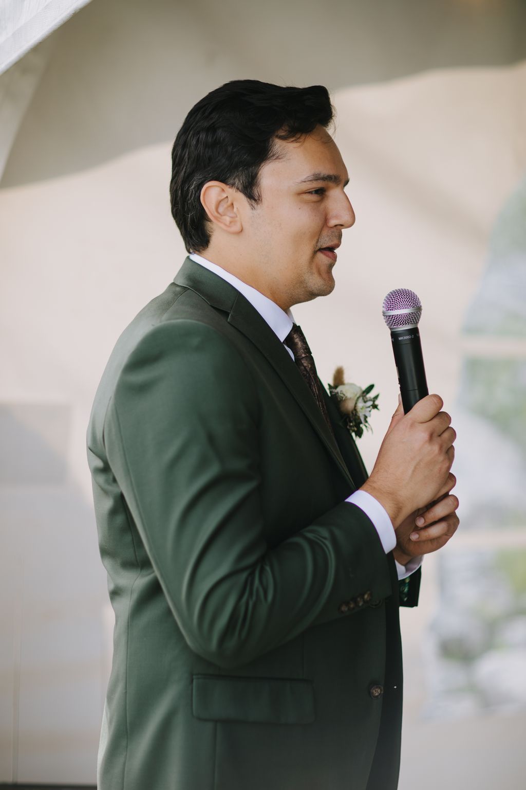 Groom in sage green suit and white shirt gazes downward, boutonniere visible, natural window light casting soft shadows
