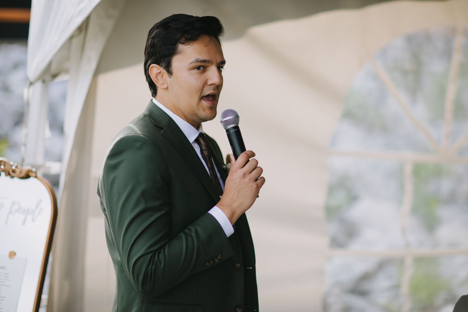 Man in dark suit and tie speaking into microphone with animated expression against soft, blurred background