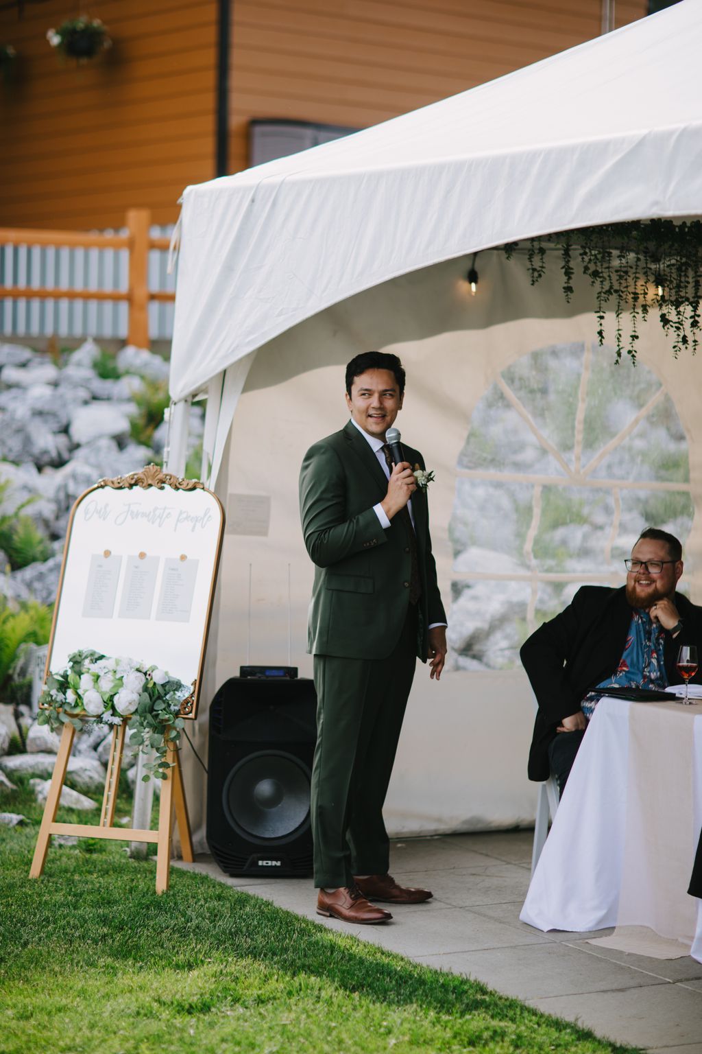 Groom in forest green suit speaking at microphone under white draped tent at outdoor wedding reception