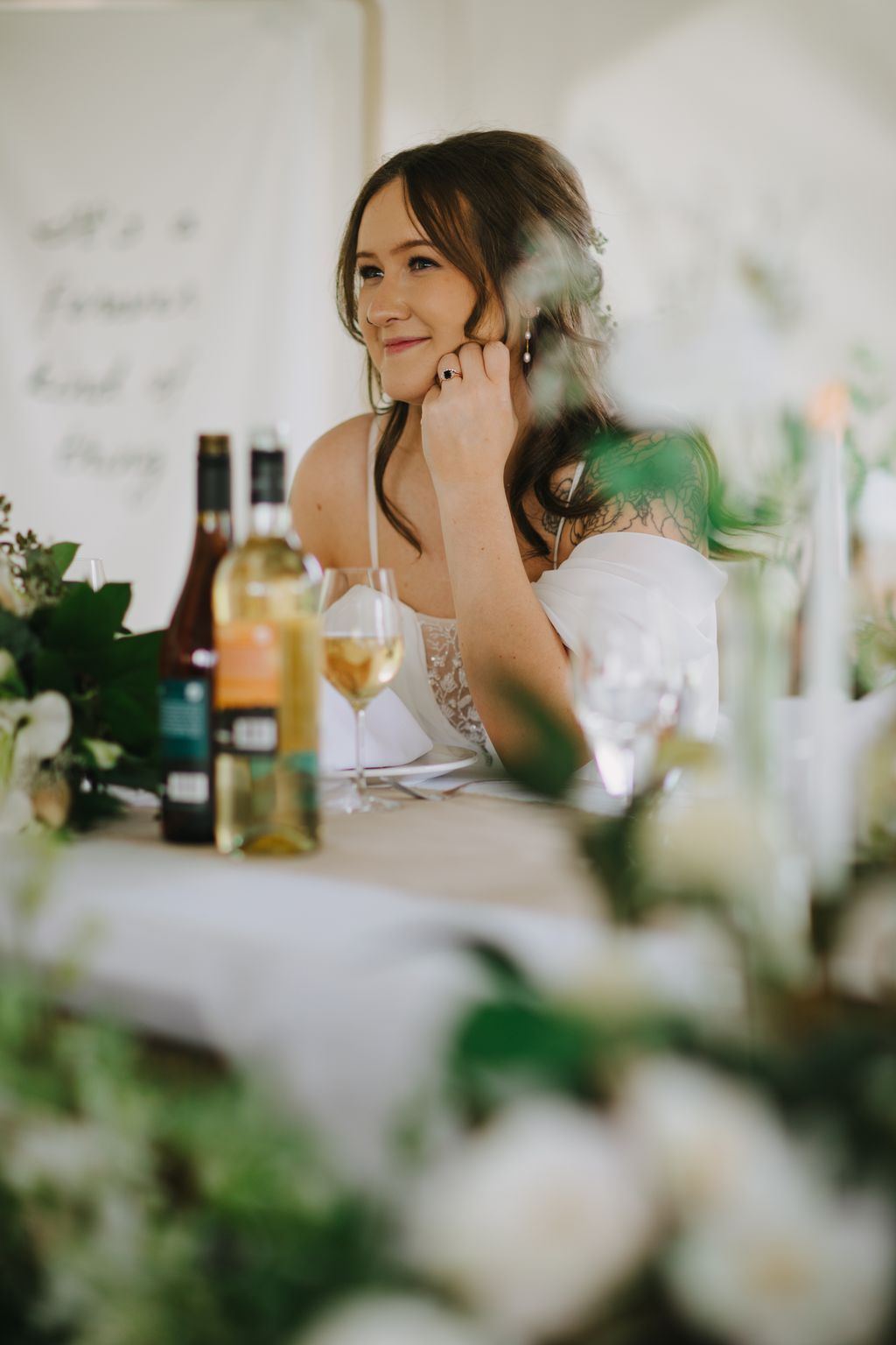 Bride with dark wavy hair and green tattoo smiling pensively at reception table with wine glasses and white flowers