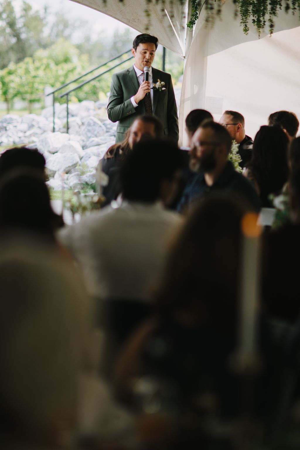Groom in sage green suit holding microphone, speaking at indoor wedding ceremony with guests in foreground and natural light from windows