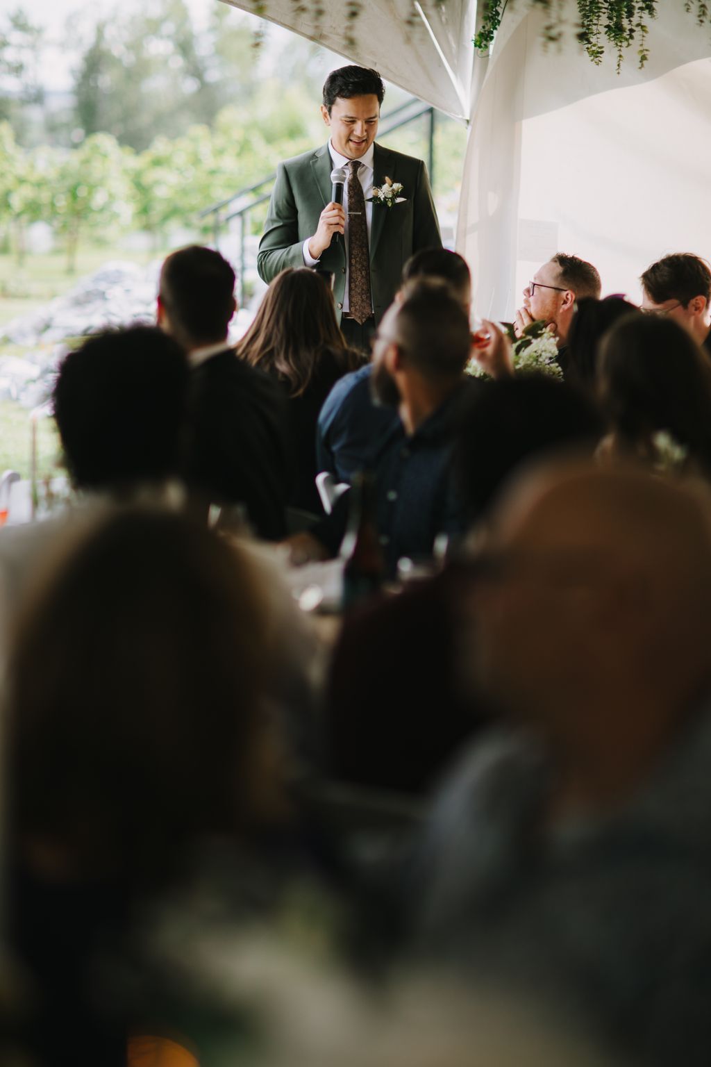 Groom speaking at microphone under white draped canopy during outdoor wedding ceremony with guests seated in foreground