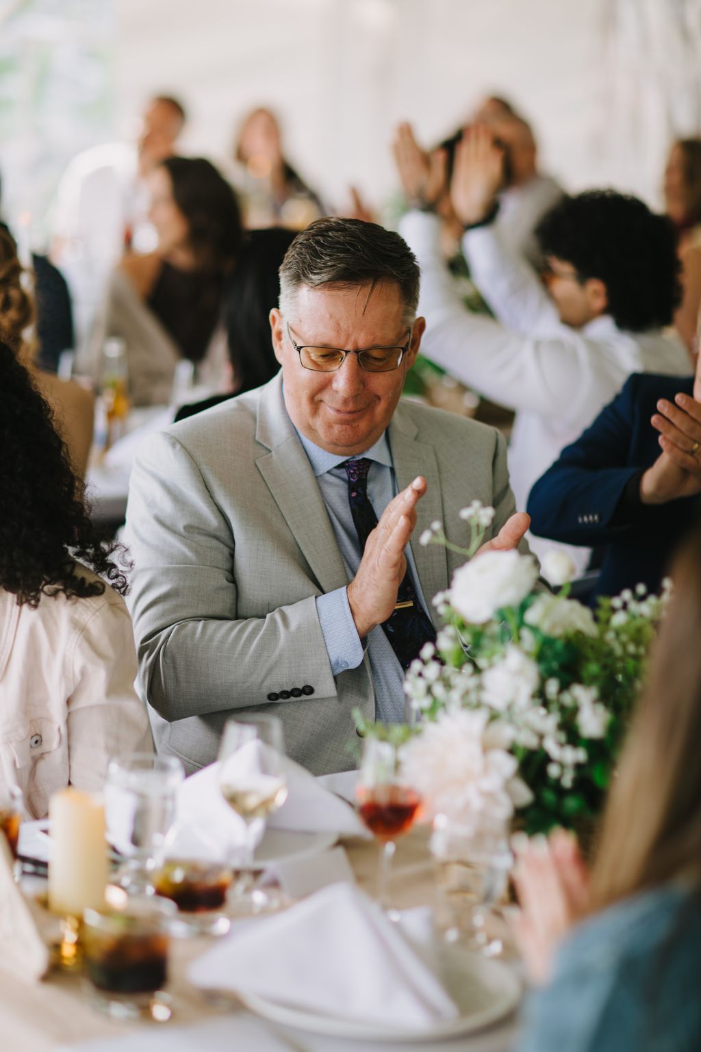 Groom in gray suit and glasses clapping during wedding reception with white floral centerpieces and guests blurred in background