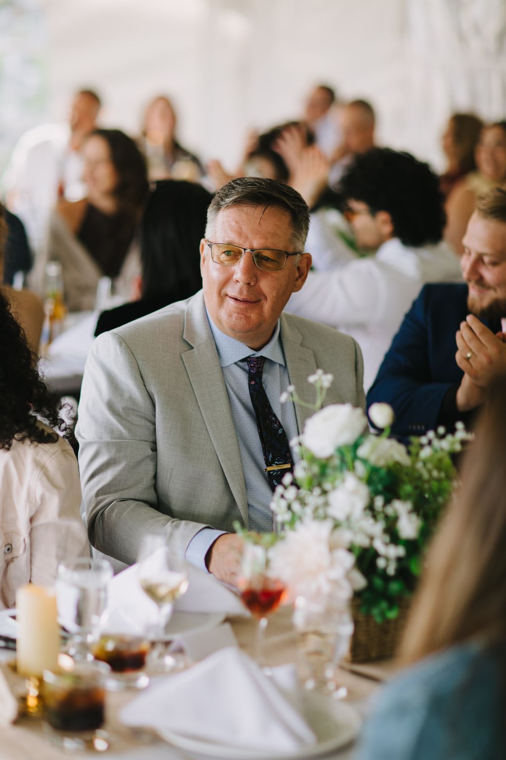 Smiling man in beige suit and glasses seated at wedding reception table with white floral arrangements and guests blurred in background