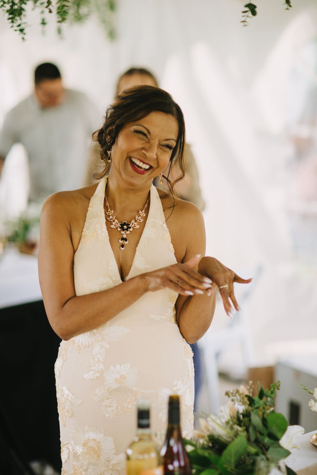 Bride in white halter dress with ornate necklace laughs joyfully, displaying wedding ring, against soft white backdrop