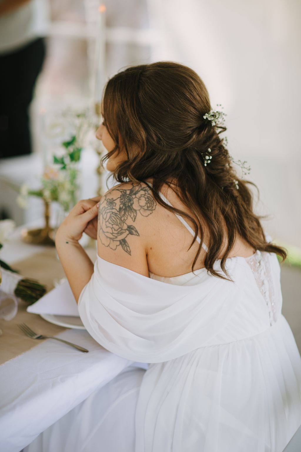 Bride with dark wavy hair and floral shoulder tattoo wearing white, adorned with baby''s breath flowers in soft natural light
