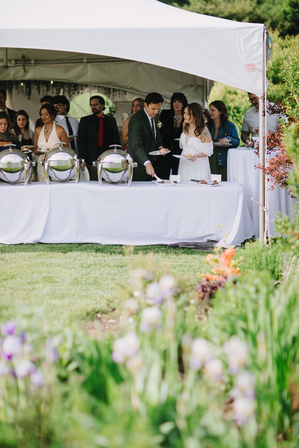 Bride and groom at outdoor wedding reception under white tent with guests, soft garden flowers in foreground