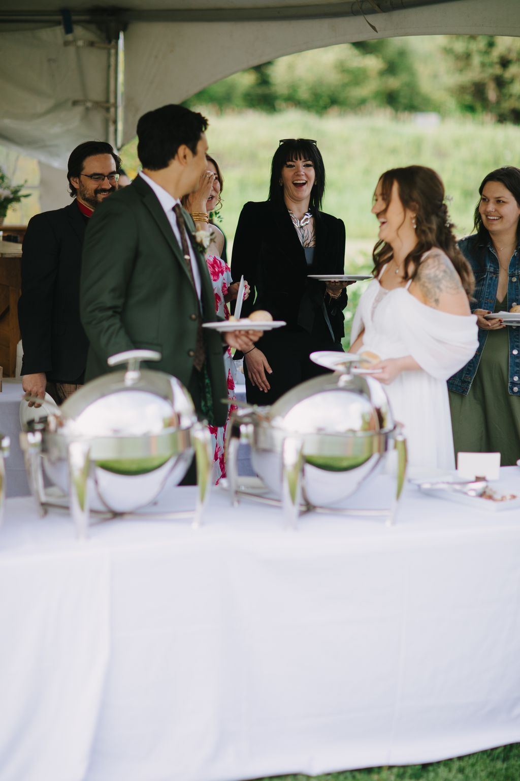 Guests laugh while holding plates at an elegant outdoor reception under a tent, with green napkins and natural daylight illuminating the celebration