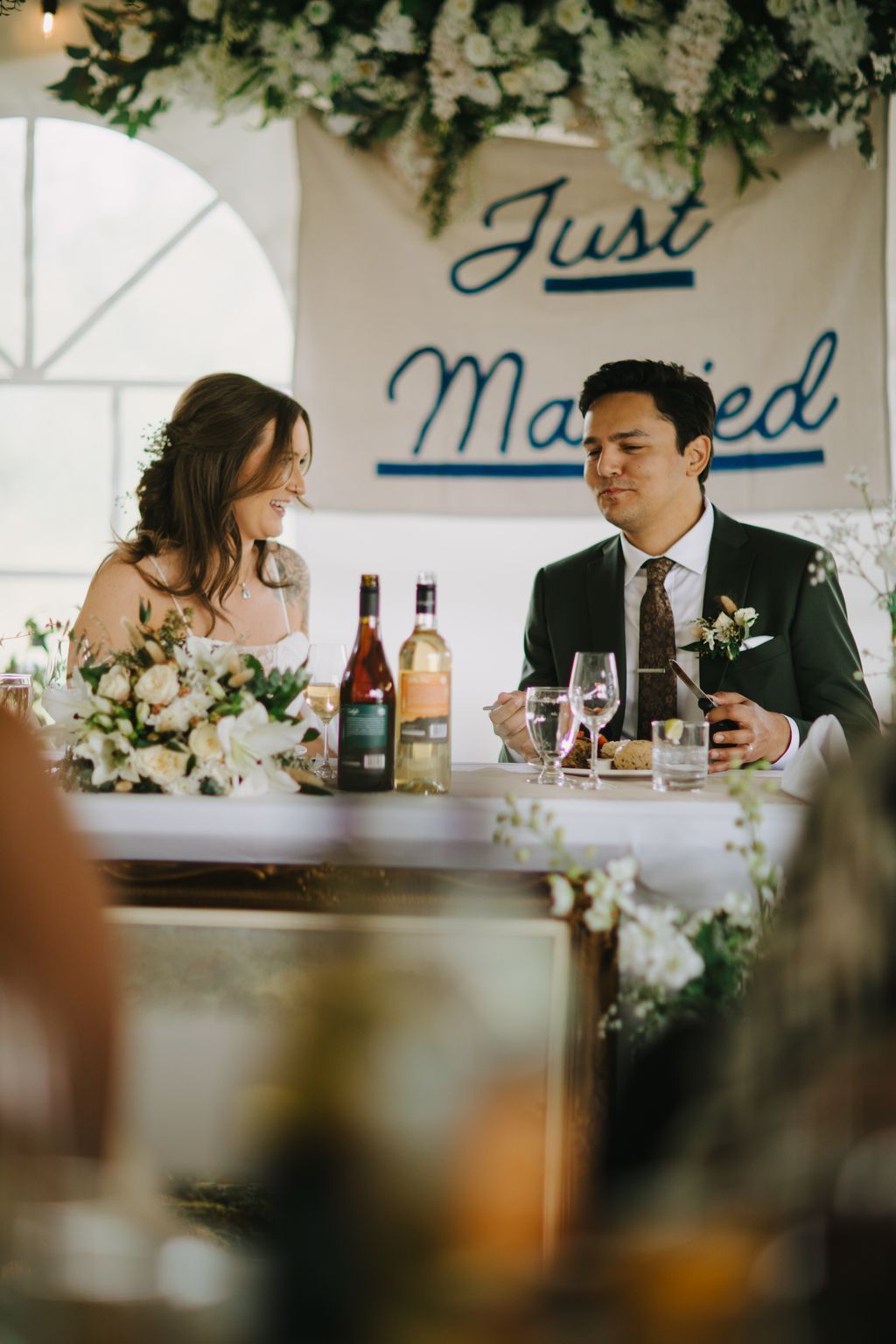 Bride and groom sit at their reception table with wine bottles and glasses, smiling beneath hanging greenery and a calligraphed sign