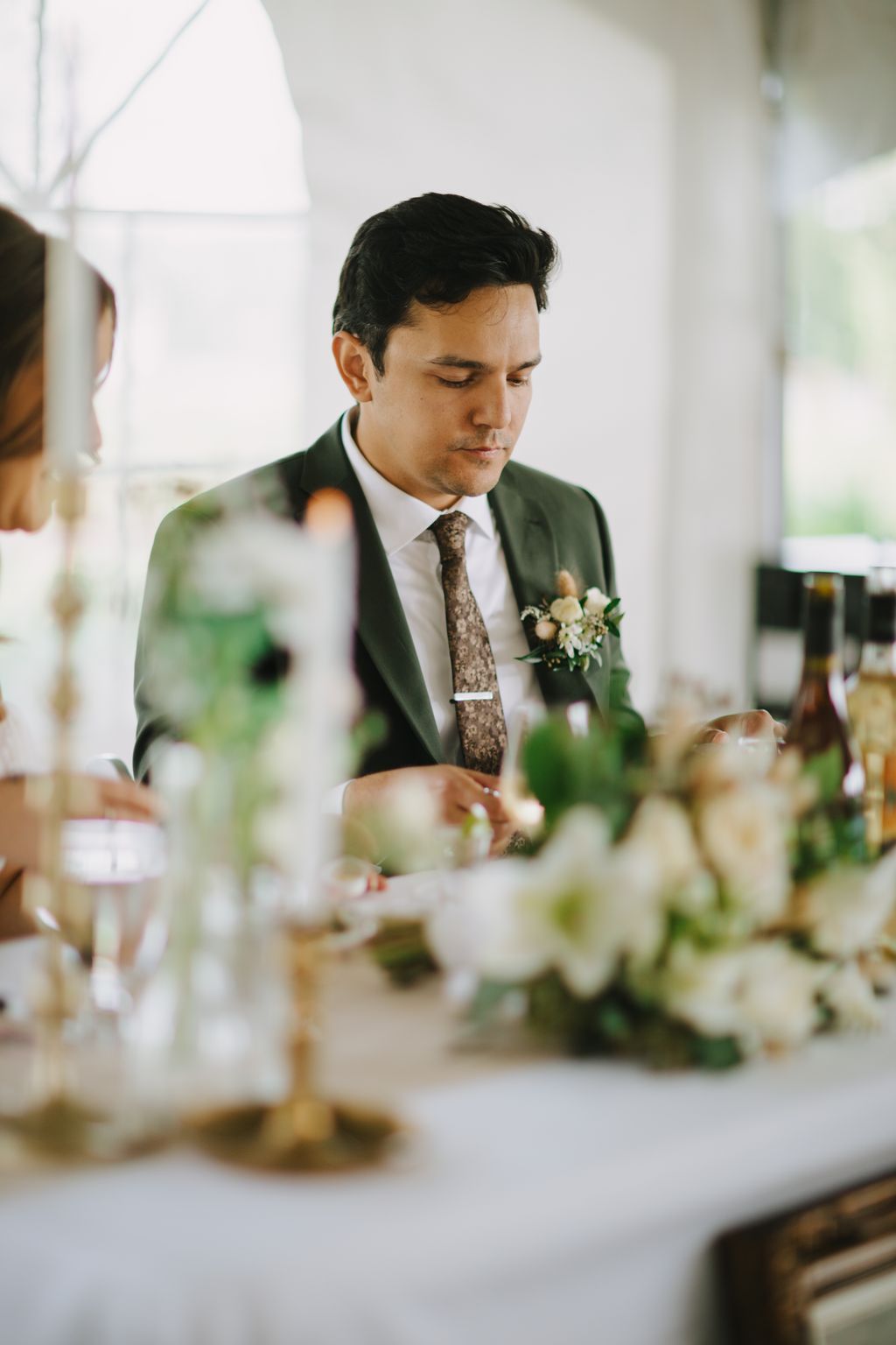 Groom in olive green suit and patterned tie with boutonniere, looking down pensively at wedding reception table with white floral arrangements