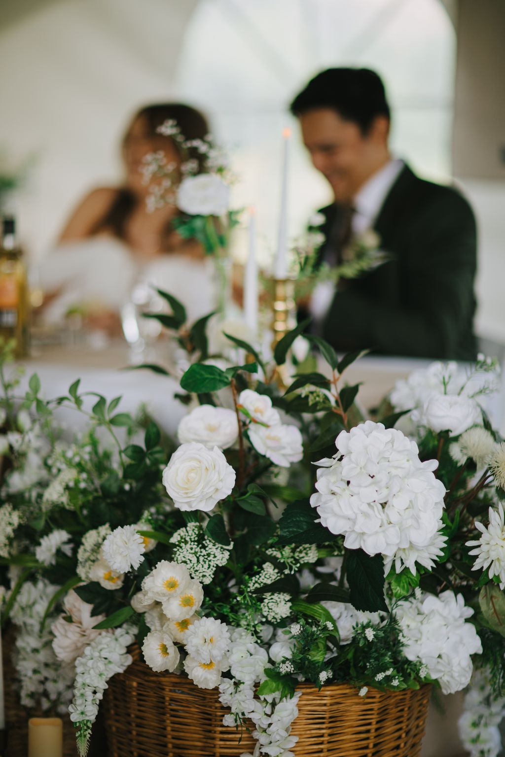 White floral arrangement in wicker basket with bride and groom softly blurred in background at wedding reception