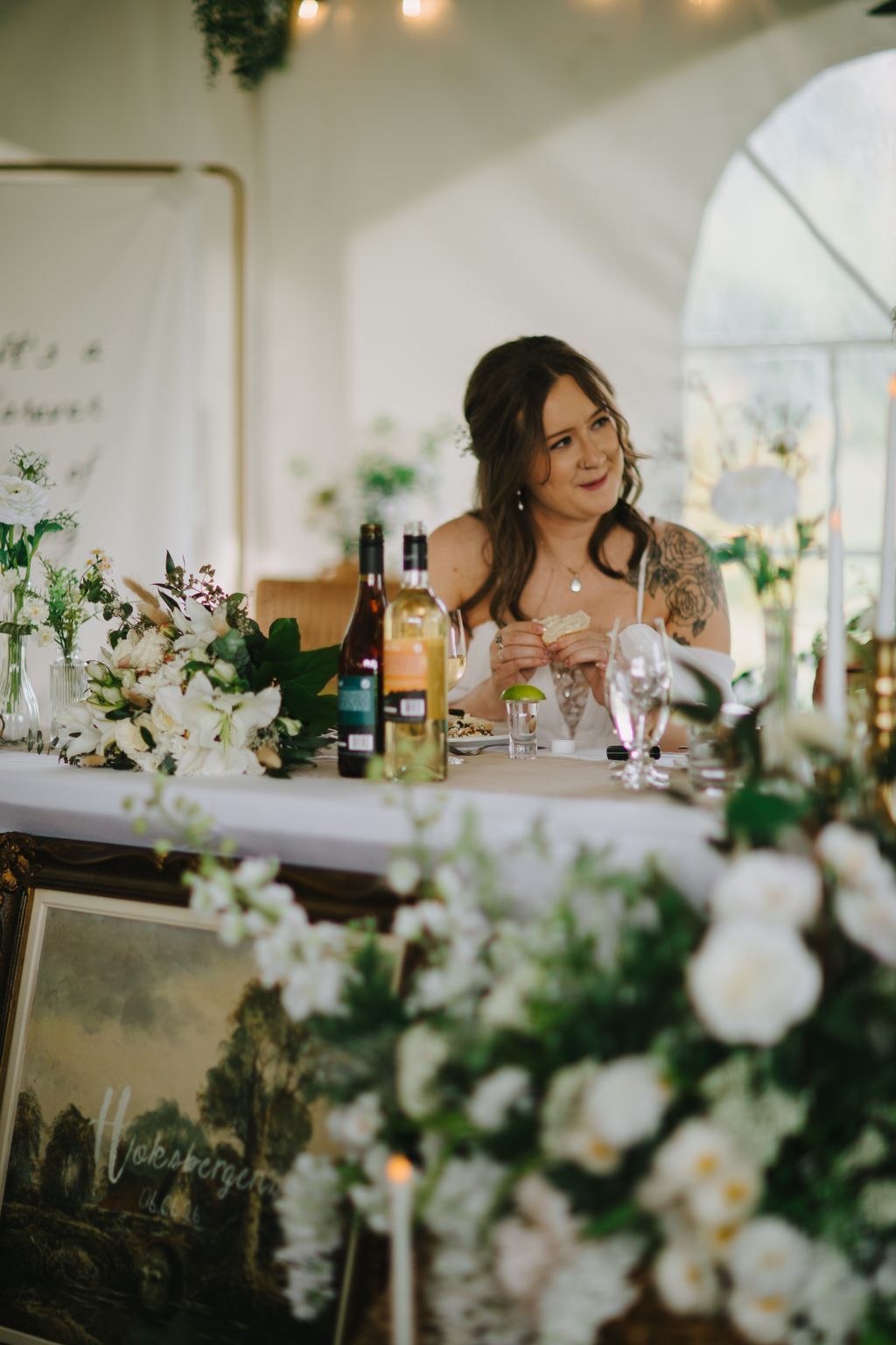 Bride in white dress seated at elegant tablescape with white florals, wine bottles, and soft overhead lighting in bright venue