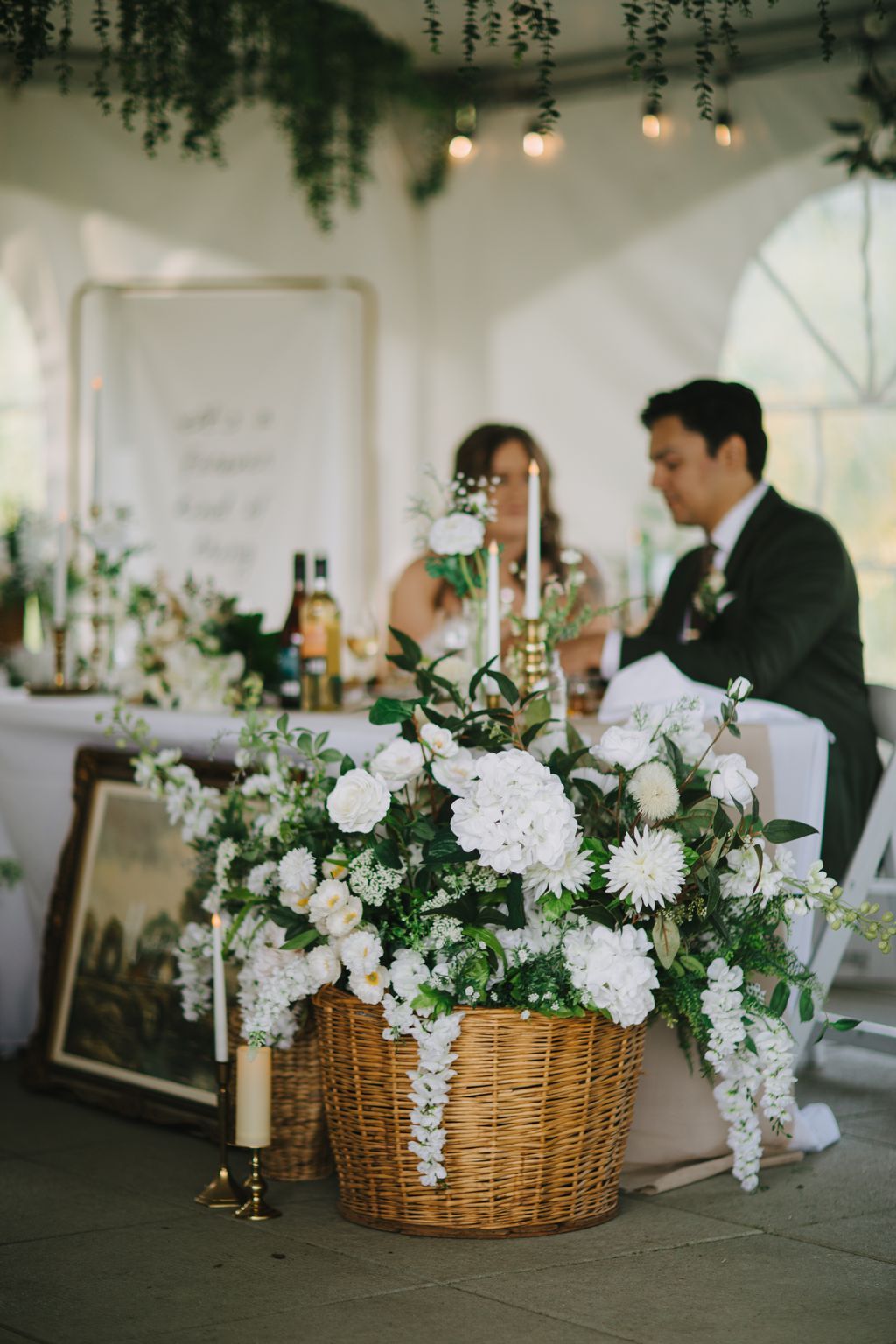 Wicker basket overflowing with white hydrangeas and daisies on an elegant reception table with hanging greenery and soft lighting above
