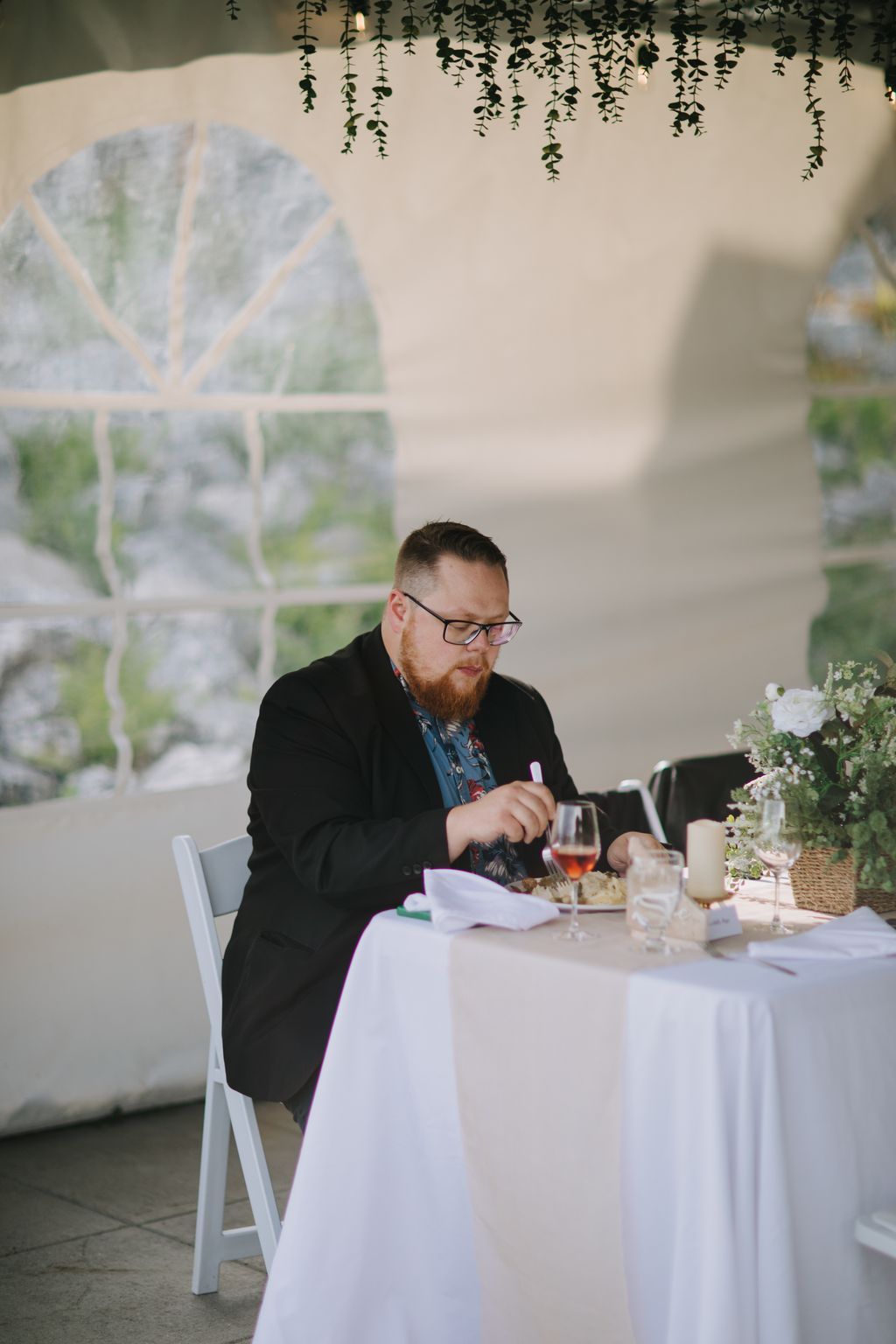 Bearded groom in glasses and black blazer enjoys a meal at white-draped table beneath hanging greenery during reception
