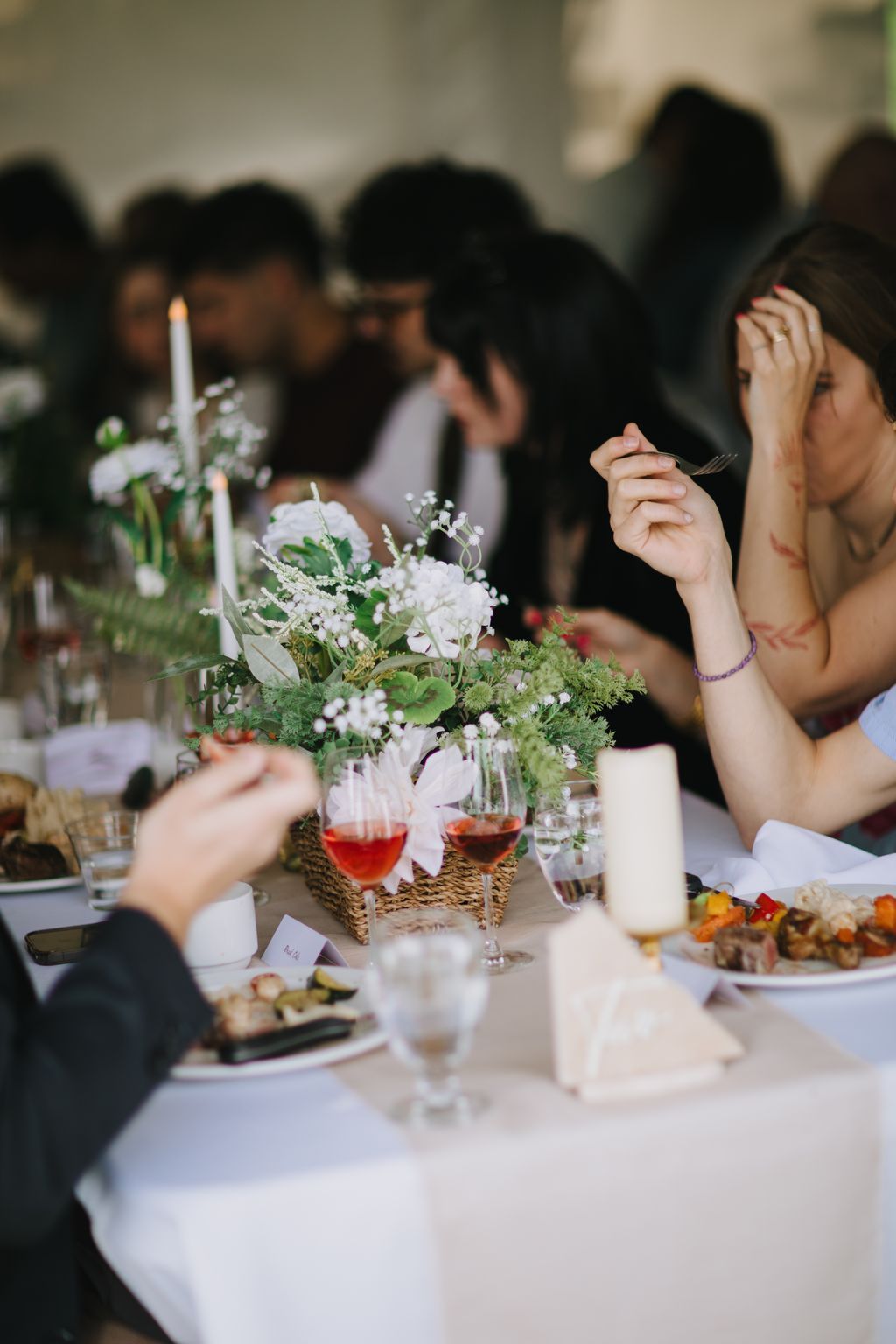 Elegant tablescape with white floral centerpiece in woven basket, red wine glasses, and blurred guests in conversation during reception