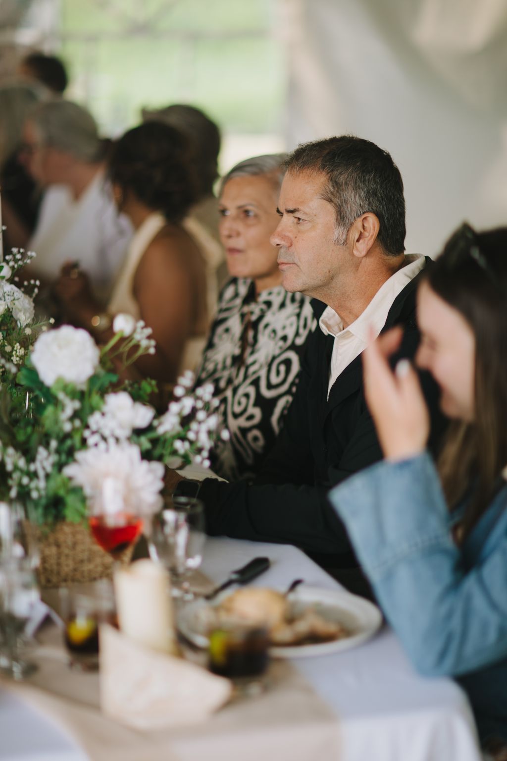 Groom in black jacket seated at wedding reception table with white floral centerpiece, gazing thoughtfully during ceremony