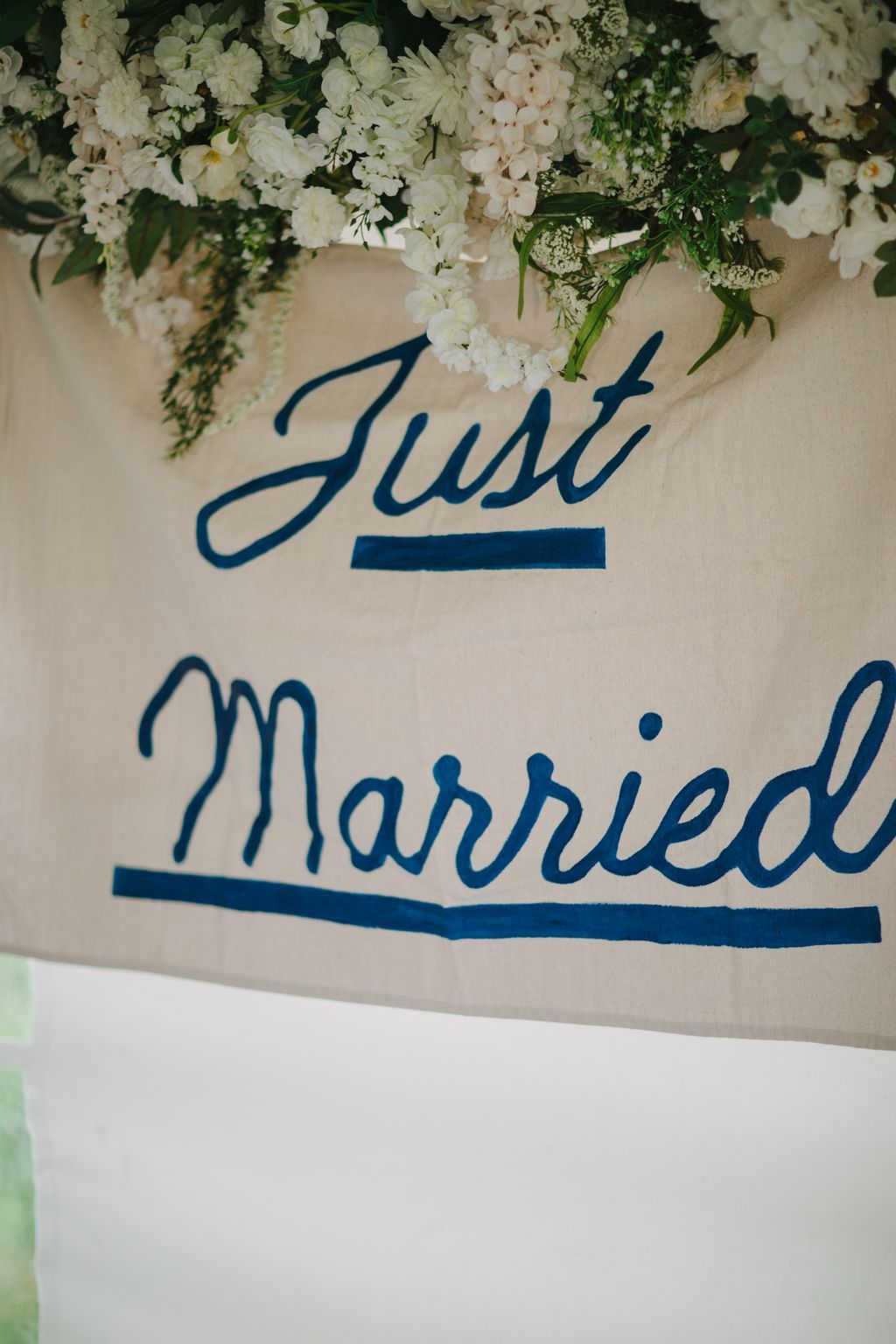 White and cream floral arrangement with lush greenery cascading above a navy calligraphy sign reading "Just Married