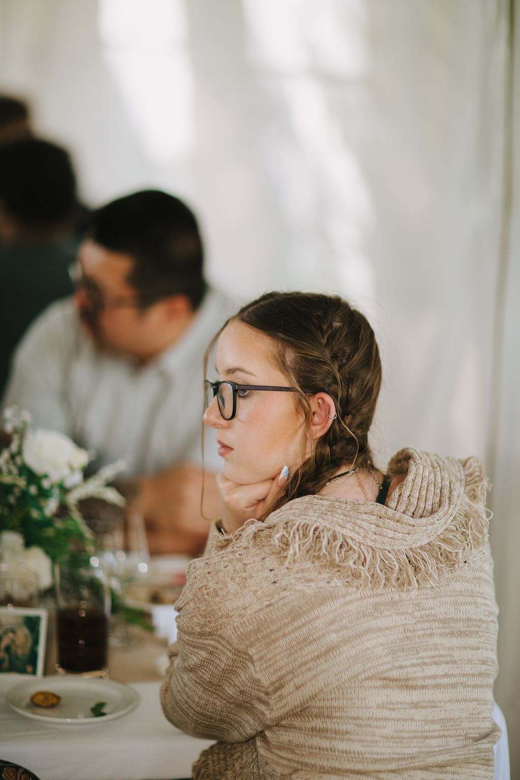 Woman wearing glasses and beige sweater looks down thoughtfully during intimate wedding moment, soft natural light