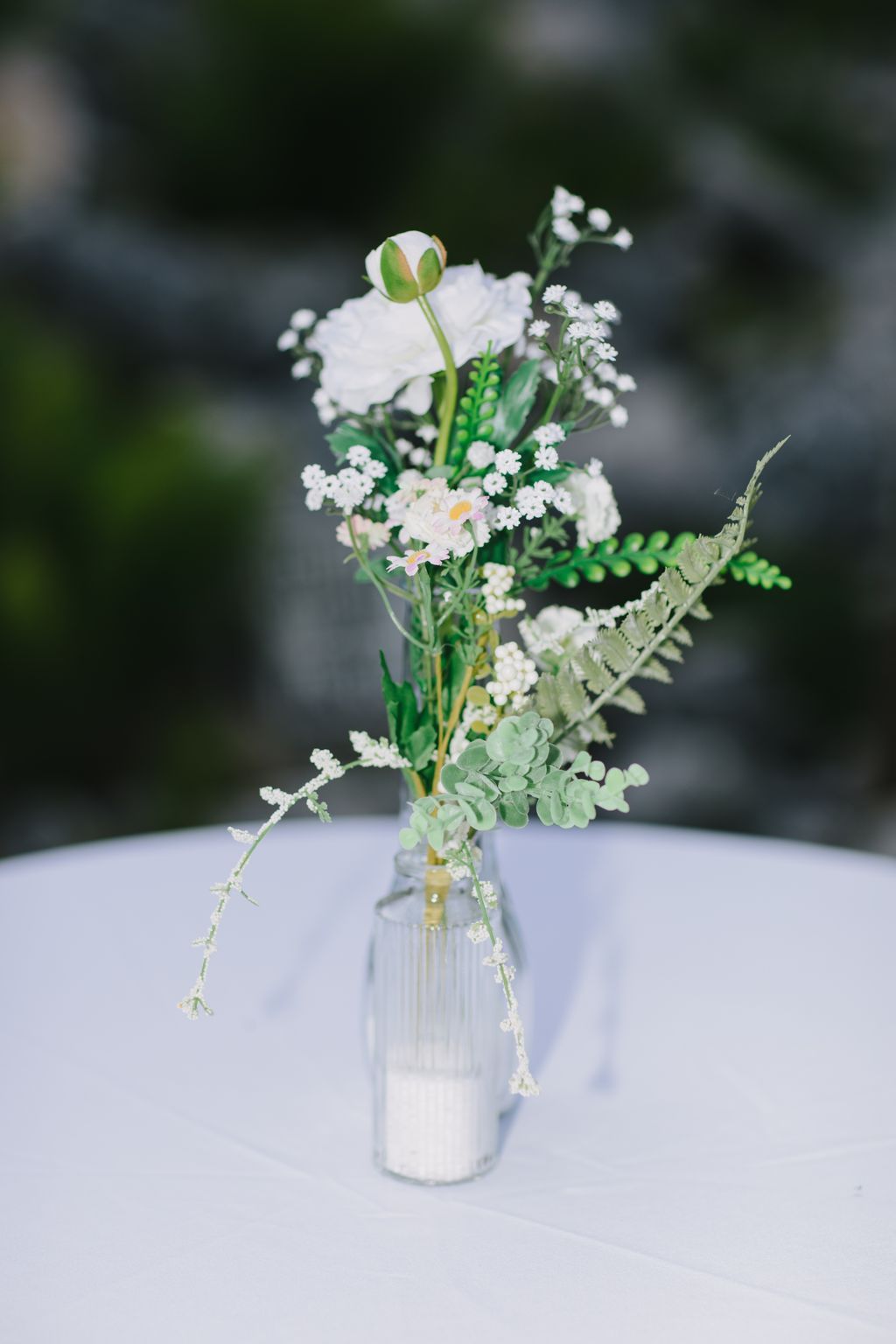 Delicate white floral arrangement with green foliage and seed pods in a clear glass vase against soft, blurred background