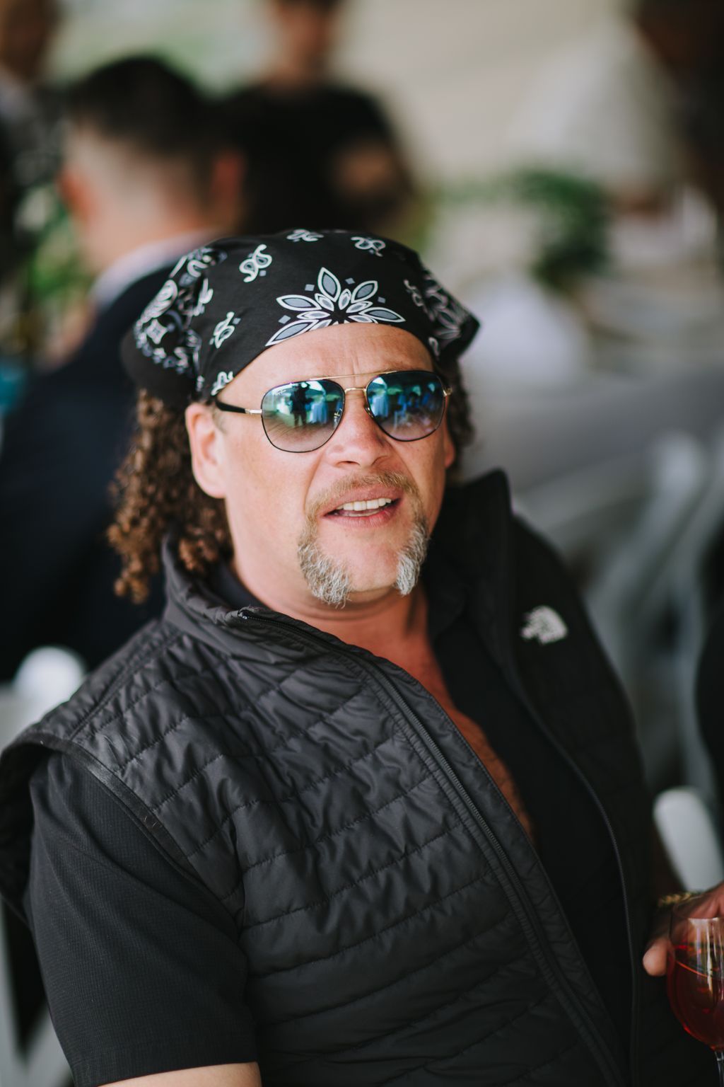 Man wearing black bandana, aviator sunglasses, and quilted jacket smiles at camera outdoors in natural light