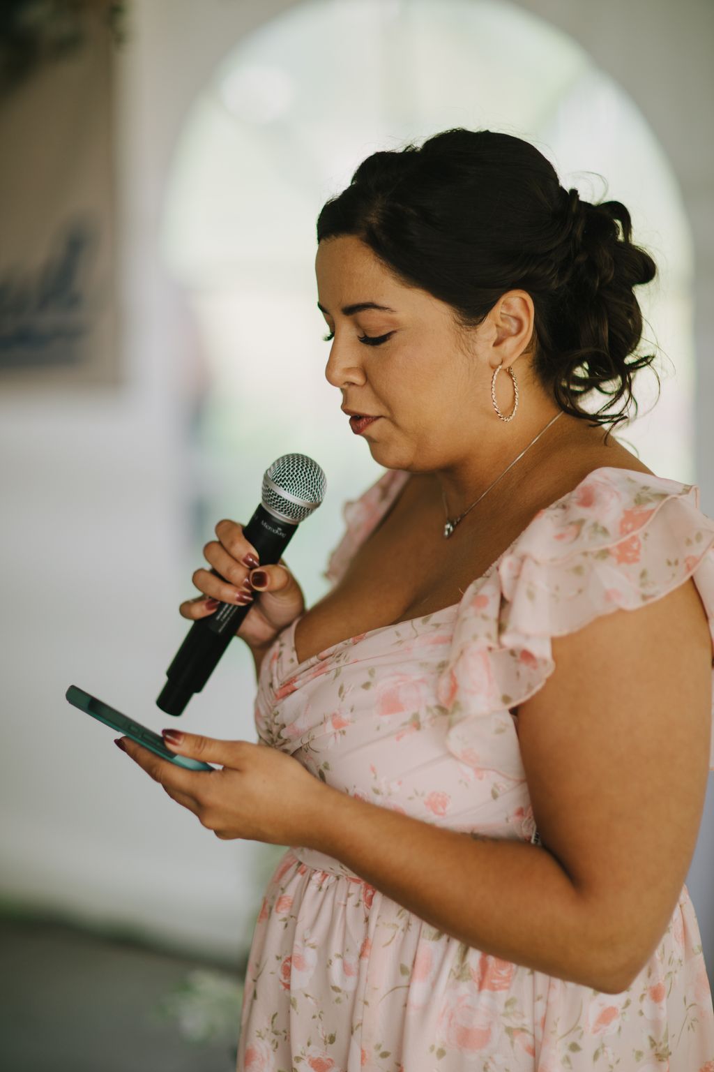Woman in pink floral dress holding microphone, eyes closed, speaking during wedding ceremony with soft natural light