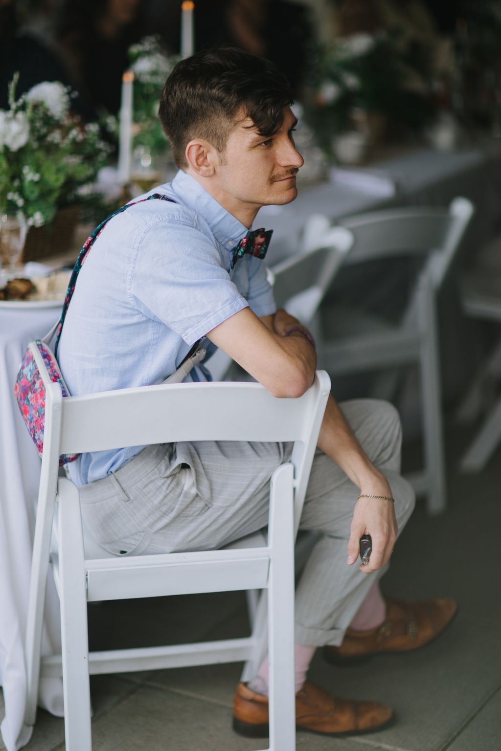 Groom in light blue shirt and floral bow tie sits thoughtfully on white chair in outdoor garden setting