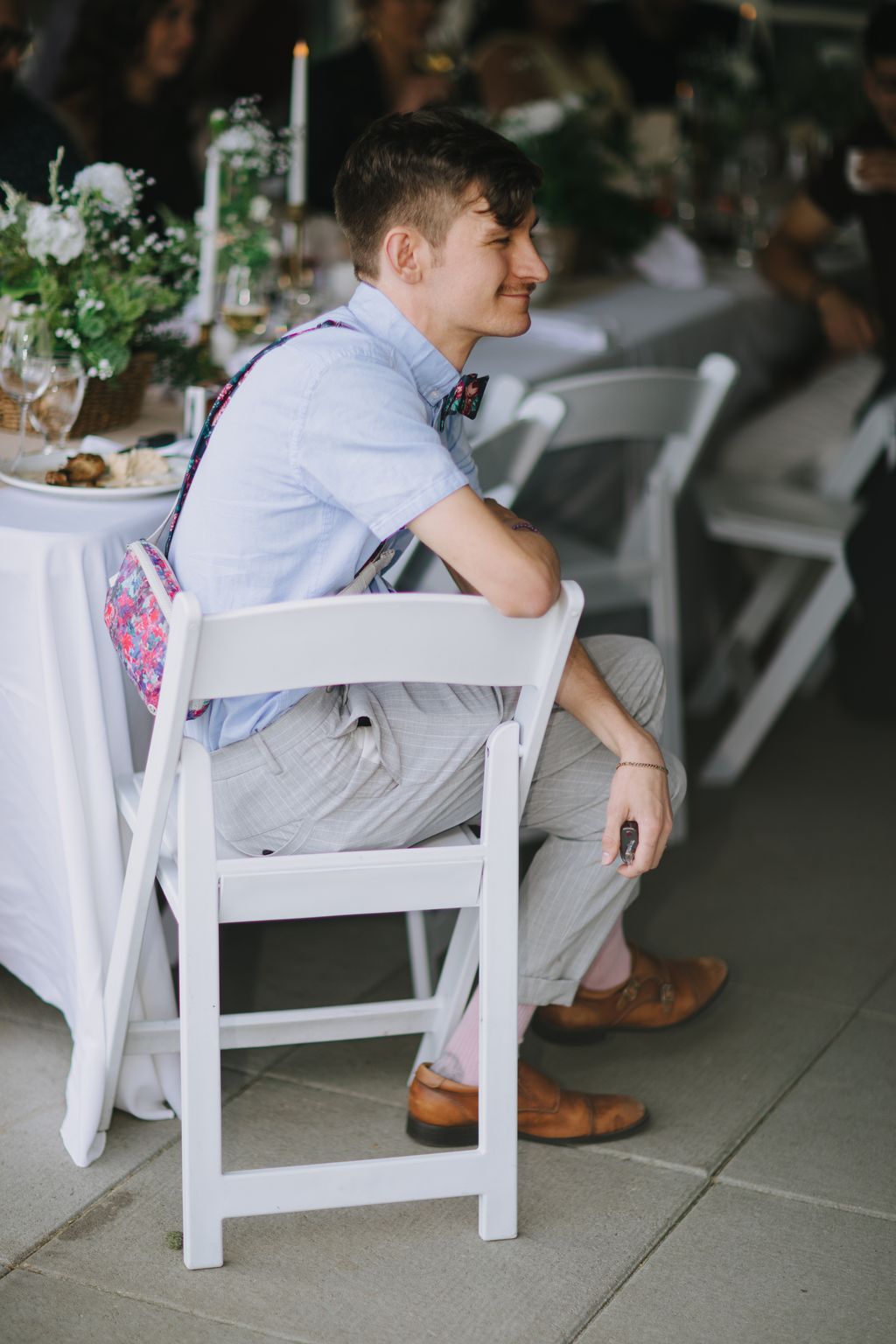 Groom in light blue shirt and floral bow tie sits smiling in profile at outdoor wedding reception
