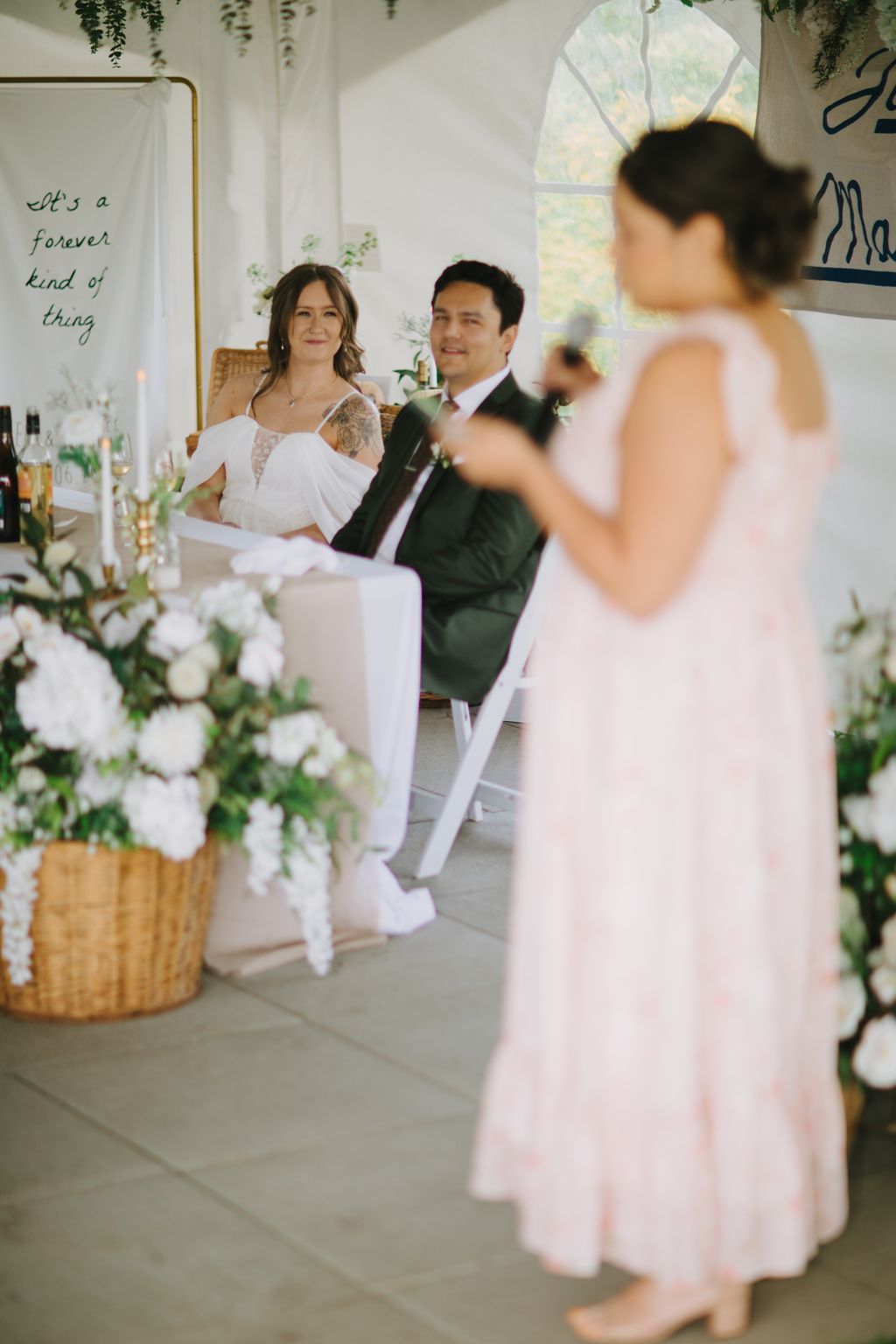 Bride and groom sit together during ceremony in bright, plant-filled venue with arched window and bridesmaid in foreground