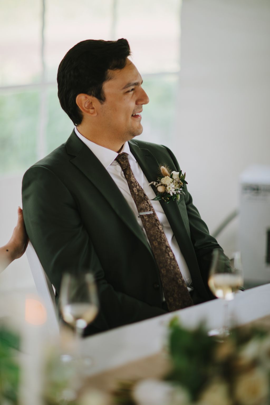 Groom in dark green suit and patterned tie smiles while wearing a delicate floral boutonniere, with soft natural light and blurred background