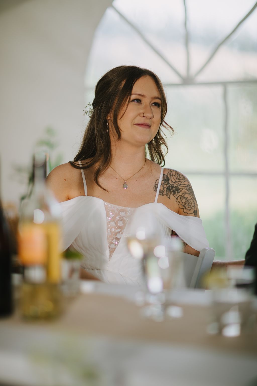 Bride in white off-shoulder lace gown with floral hair piece gazes thoughtfully in bright, modern venue