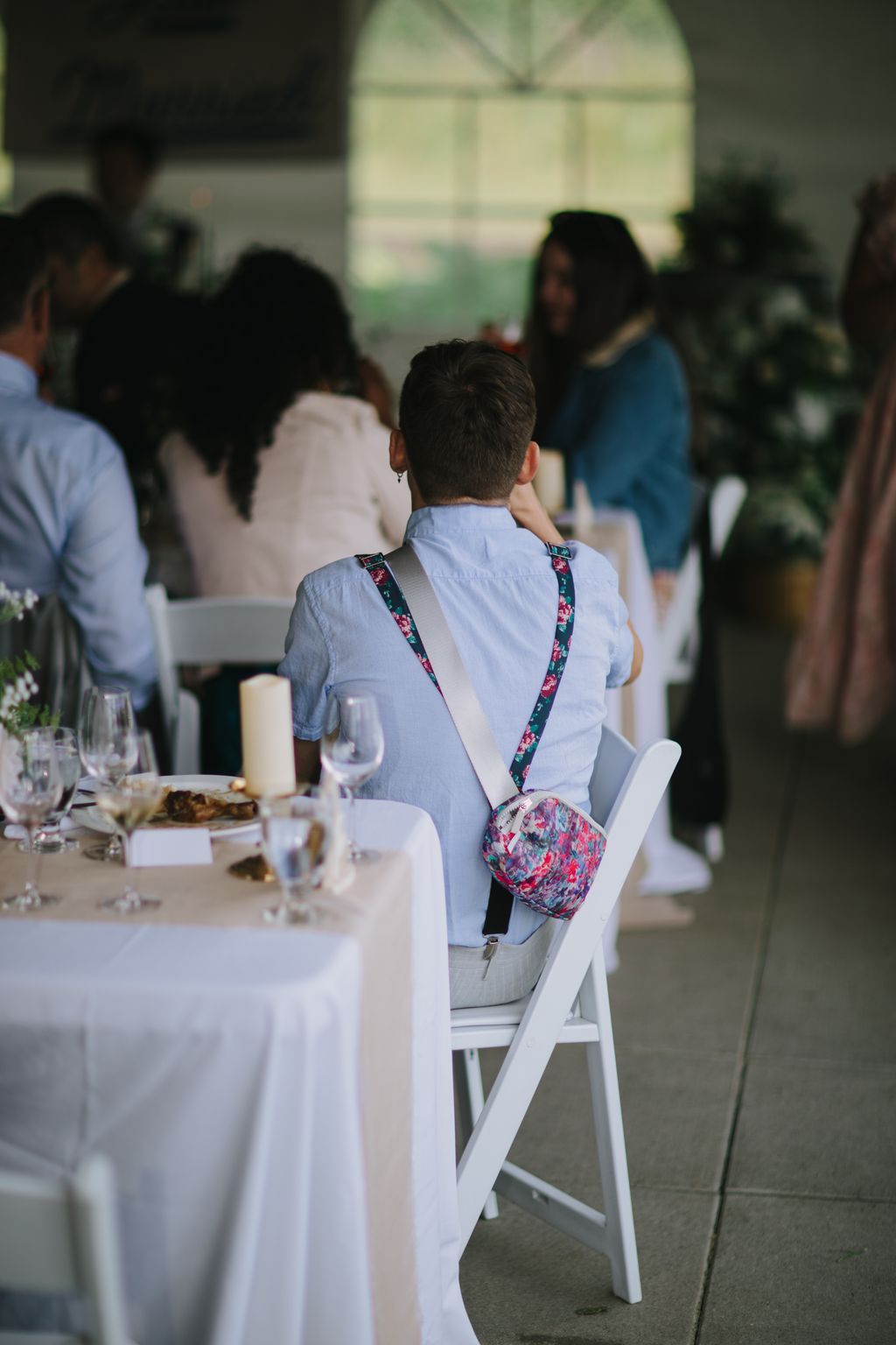 Young boy in light blue shirt with floral crossbody bag sits at white-clothed reception table during wedding celebration