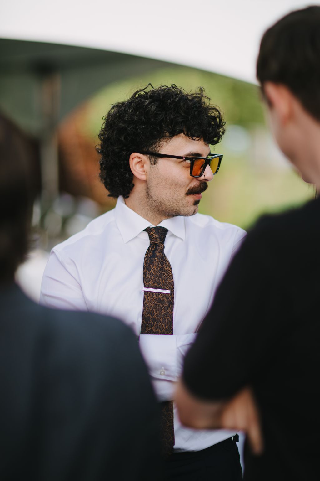 Groom in white shirt and patterned tie wearing sunglasses, photographed outdoors with soft natural light and blurred green background
