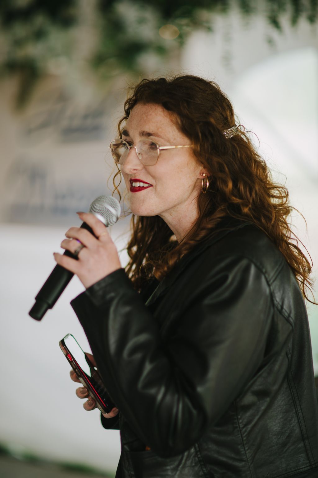 Woman in glasses and black jacket speaking into microphone at outdoor wedding event with blurred green foliage background