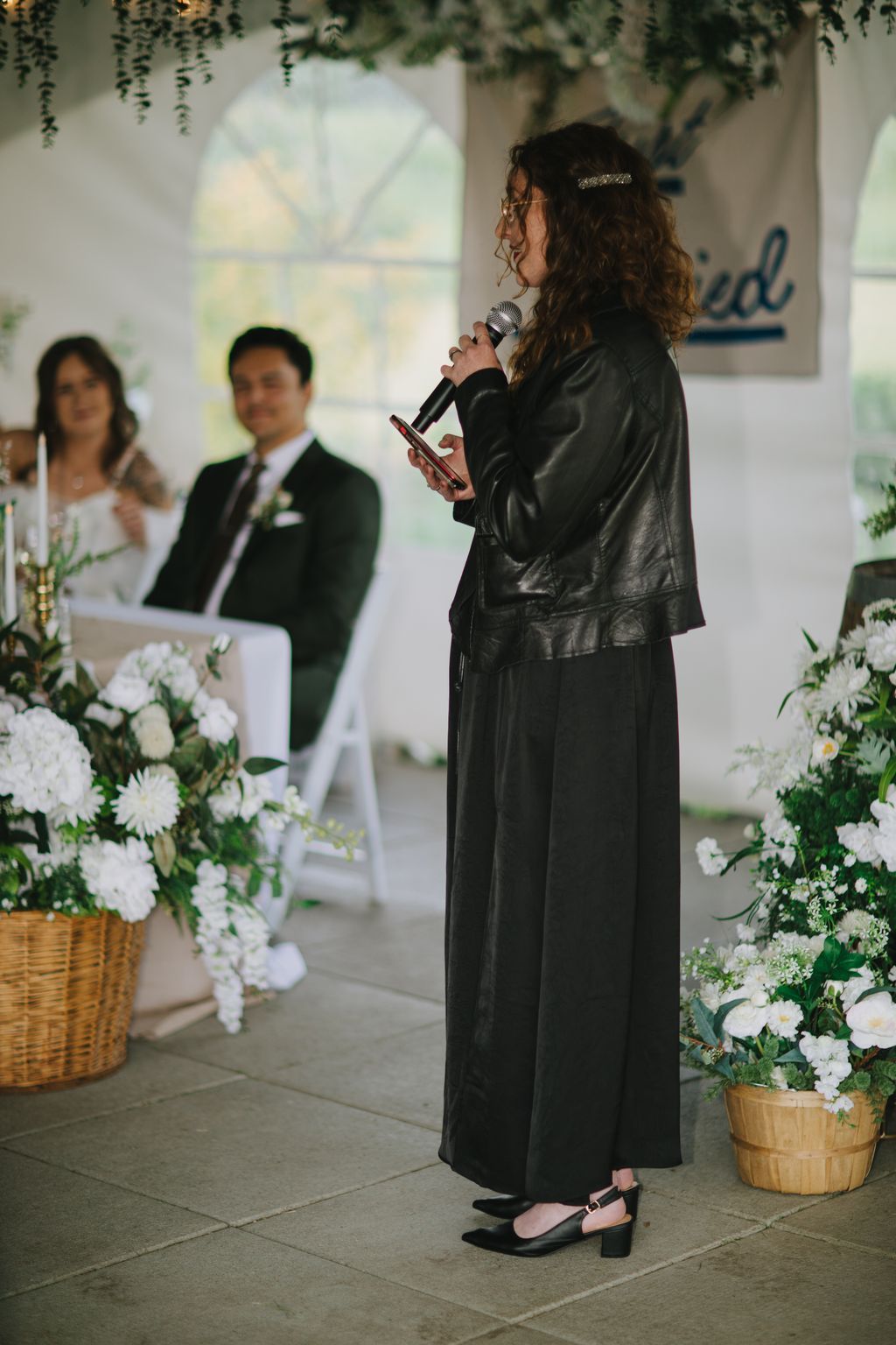 Woman in black leather jacket speaking at microphone during indoor wedding ceremony with large windows and greenery overhead
