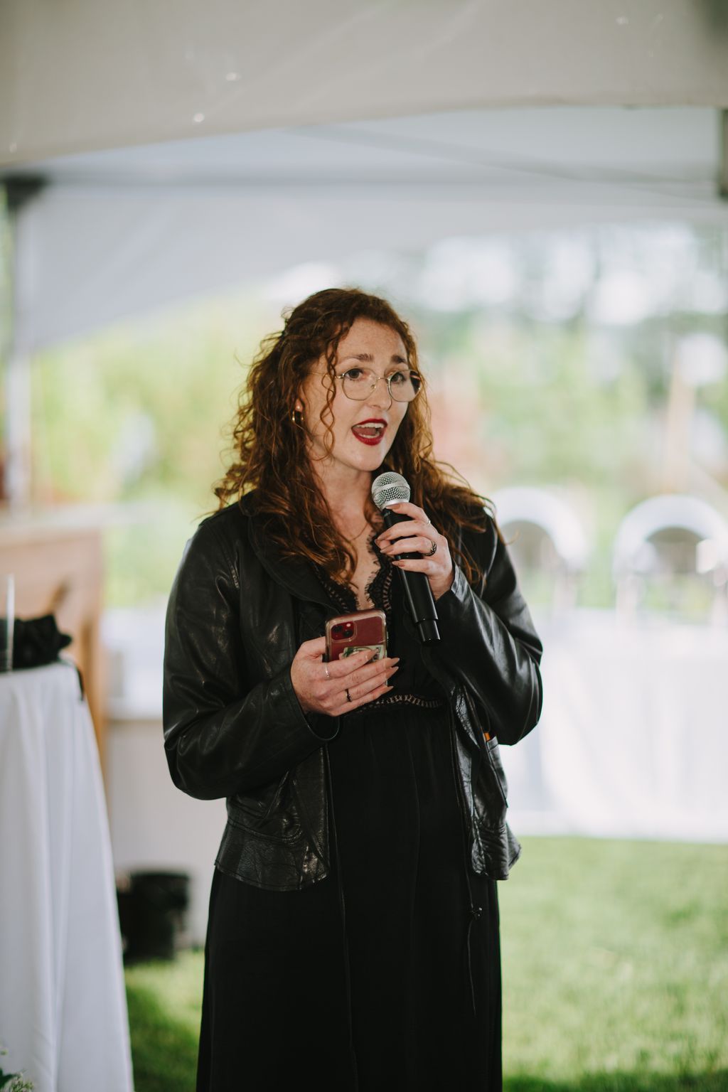 Woman with curly brown hair wearing glasses and red lipstick speaks into microphone at indoor event, holding phone