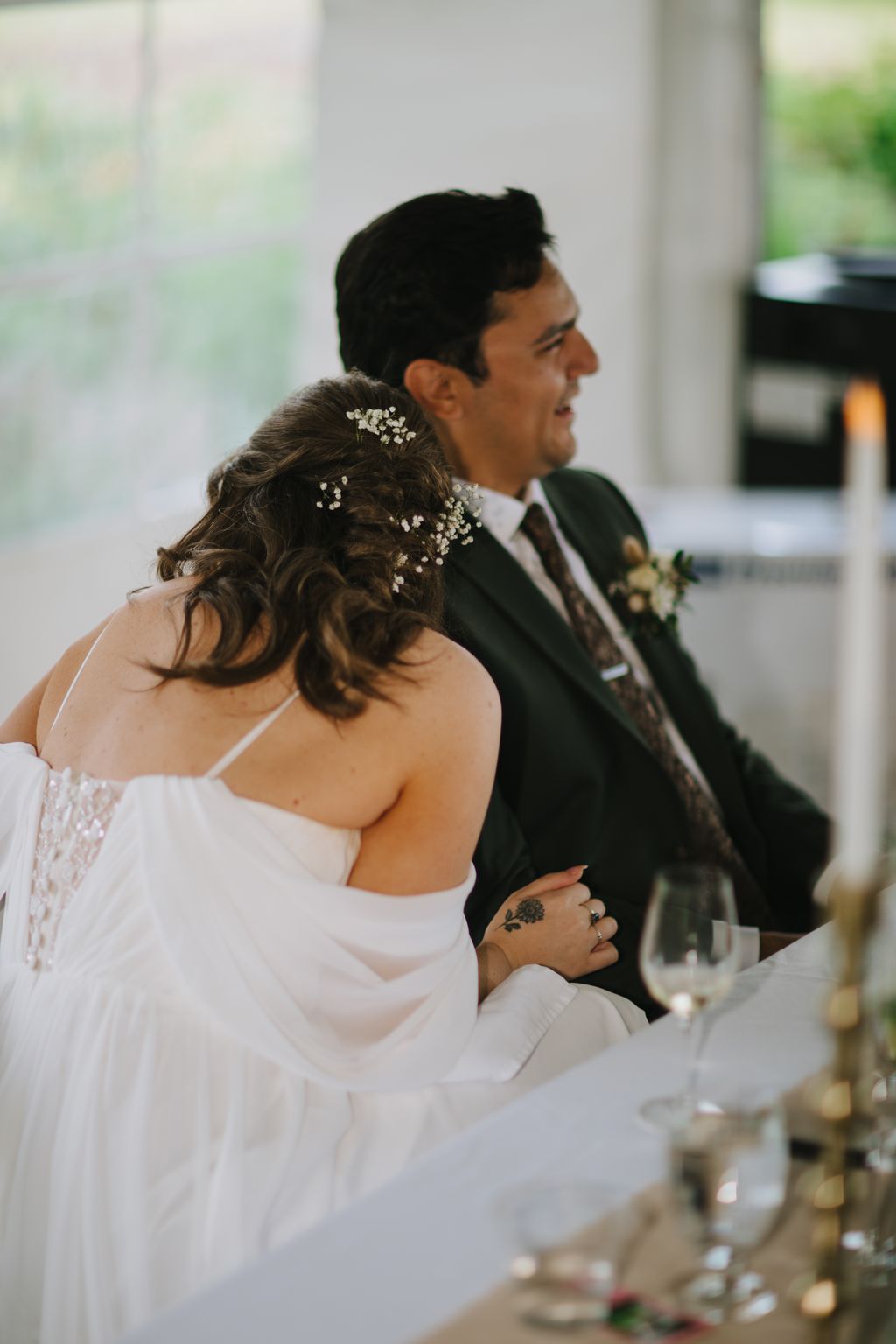 Bride and groom in profile, she in white dress with baby''s breath in her hair, he in green suit, smiling during ceremony with soft natural light