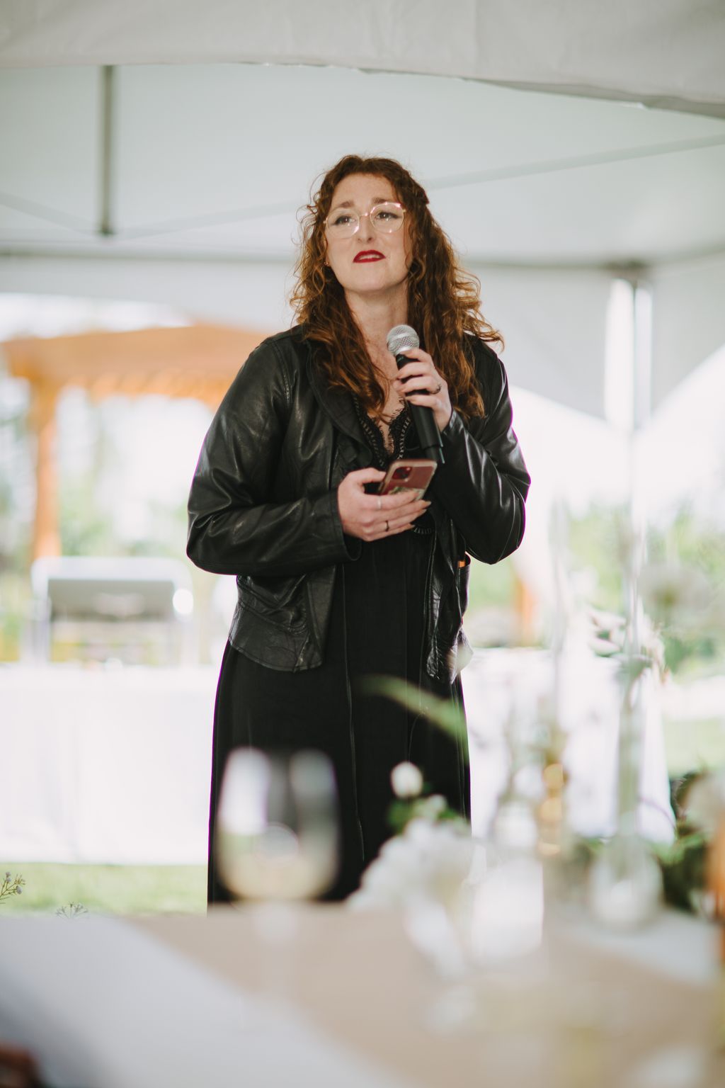 Woman with curly auburn hair wearing glasses and black leather jacket speaks into microphone at indoor event with blurred background