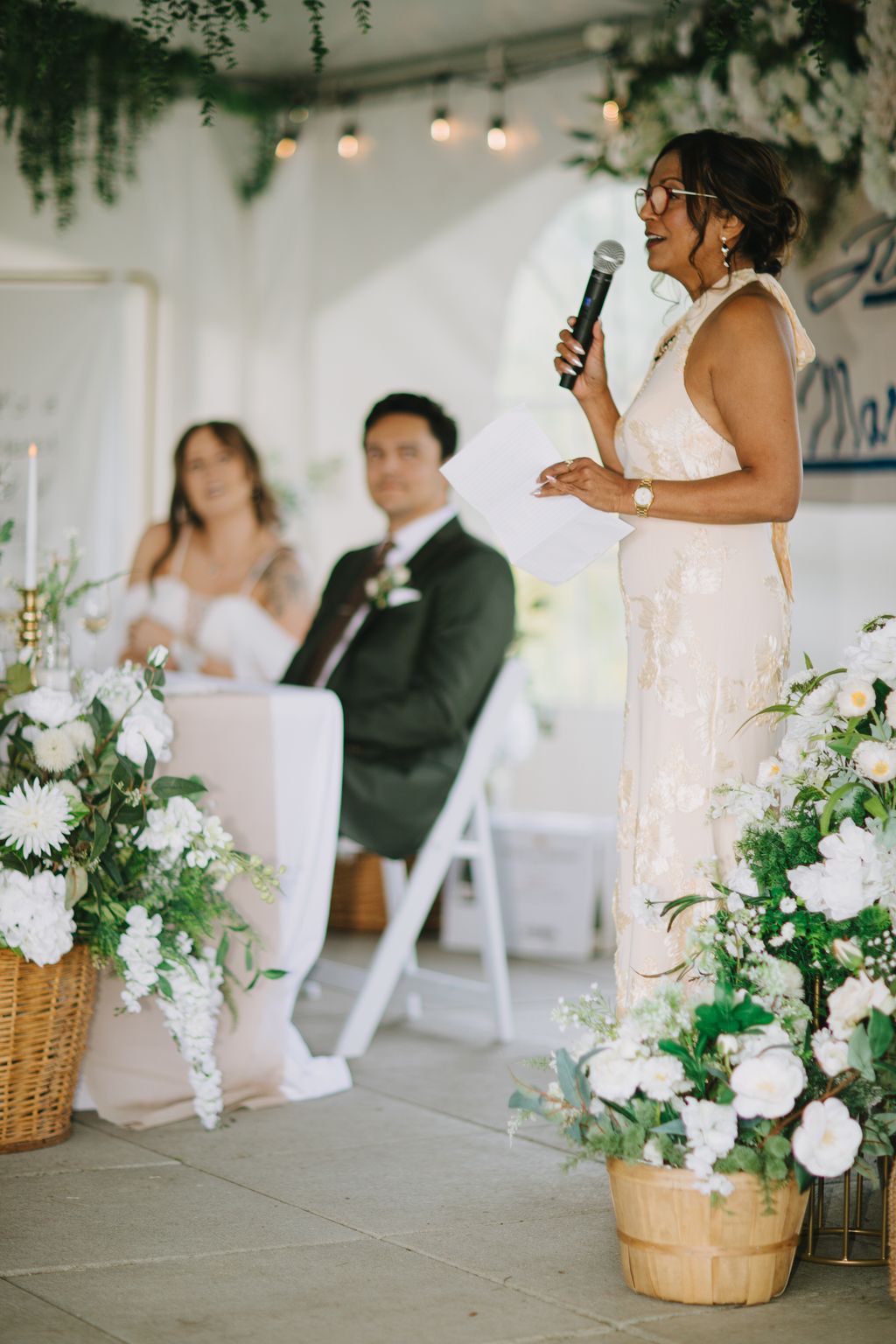 Woman in cream dress speaking at microphone during elegant indoor wedding ceremony with greenery, hanging lights, and blurred groom in background