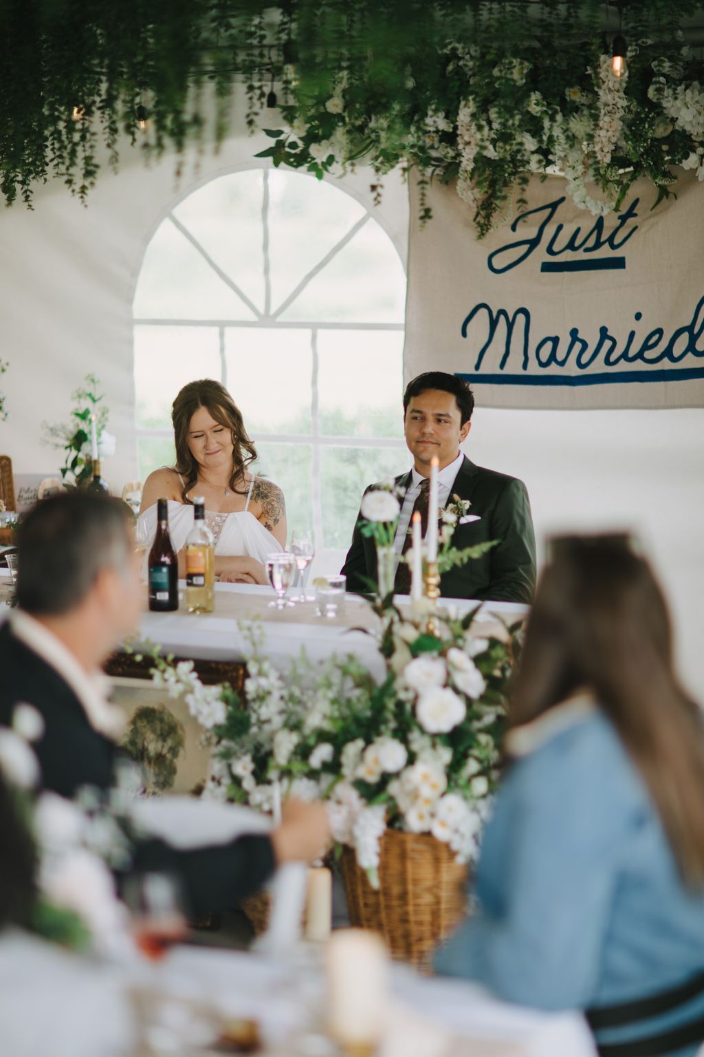 Bride and groom sit at reception table beneath hanging greenery, with white flowers and soft natural light from arched window behind them