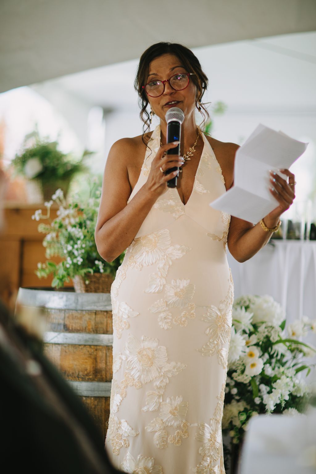 Bride in cream embroidered halter gown speaks into microphone wearing glasses, soft natural light and blurred greenery behind her