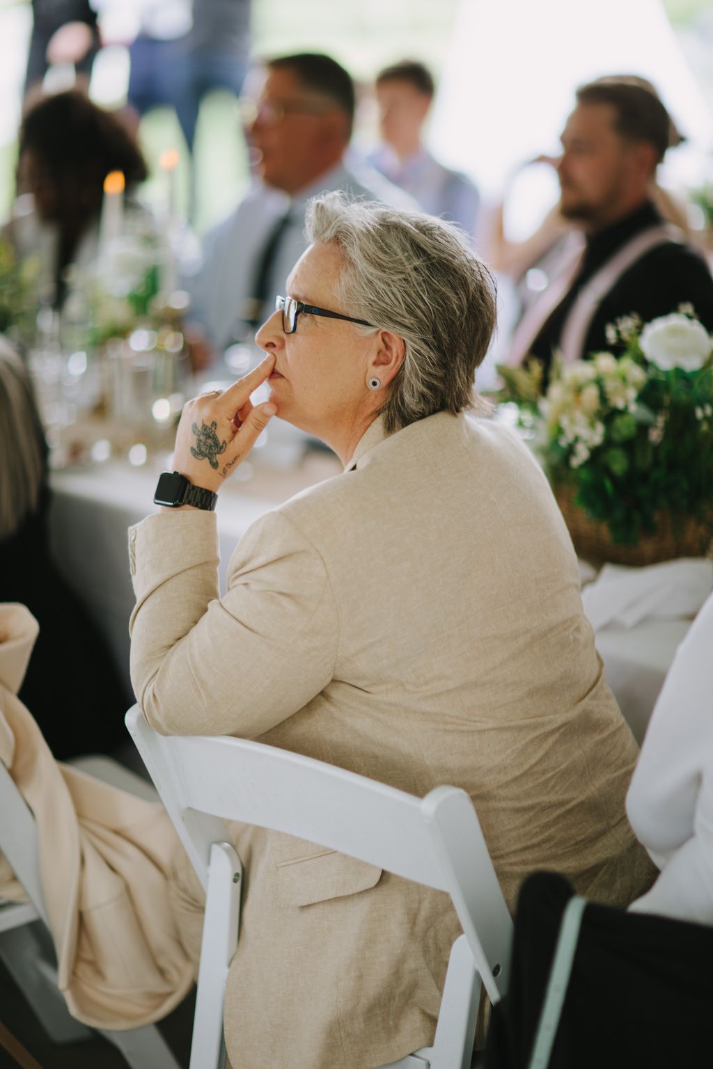 Thoughtful guest in beige blazer and glasses touches chin during wedding ceremony, with blurred attendees in background