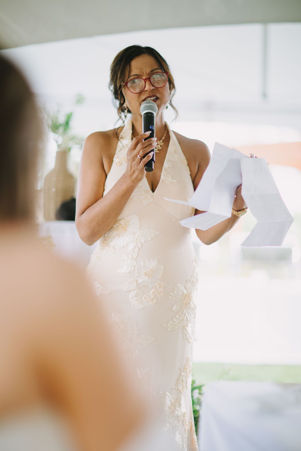 Bride in embellished ivory halter gown speaking into microphone at wedding reception, wearing red glasses and pearl necklace