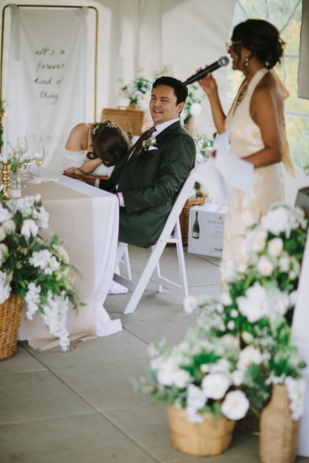 Groom in sage green suit smiles while holding bride during indoor ceremony with white florals and natural light