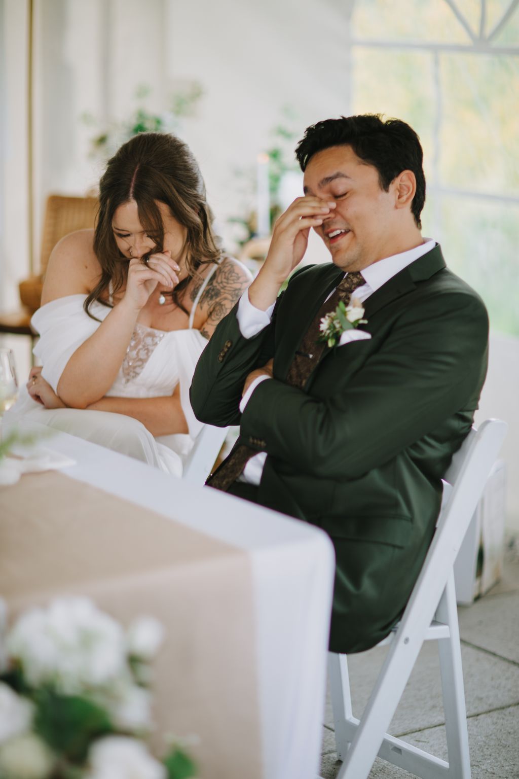 Bride and groom laugh together during ceremony, groom wiping away tears of joy, soft natural light and greenery in background