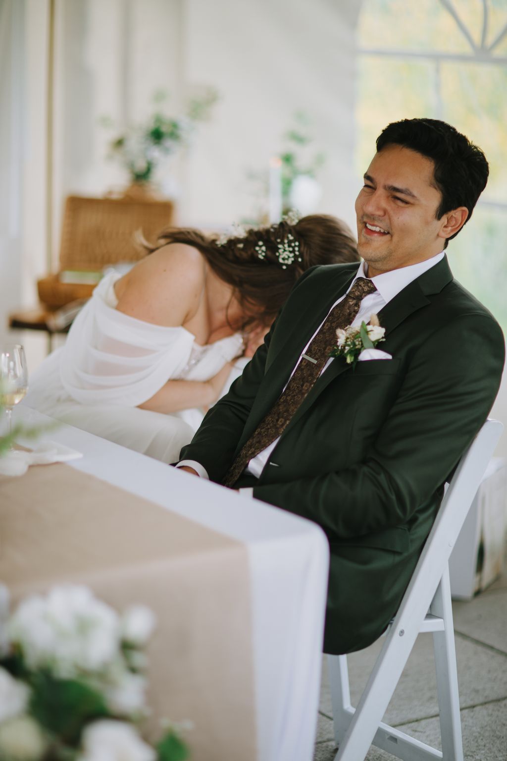 Bride and groom smile during ceremony, bride wearing white dress with baby''s breath in hair, groom in dark suit with patterned tie, holding ceremony program