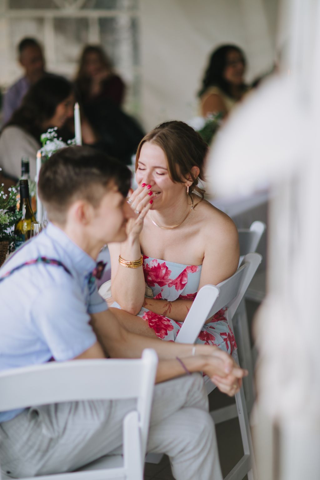 Woman in floral strapless dress laughing with hand to face during wedding reception, seated among guests
